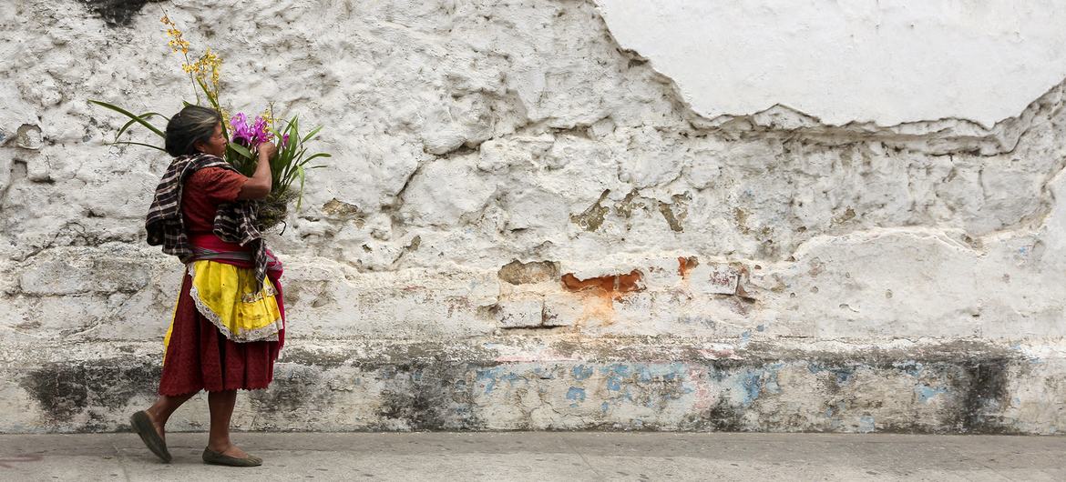 زنی سبدی از گل در کنار خیابان های سنگفرش شده و دیوارهای فرسوده در آنتیگوا، گواتمالا در دست دارد.